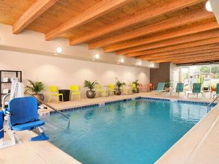 An accessible hotel indoor swimming pool with a wooden ceiling, blue pool lift chair, and bright yellow accent chairs